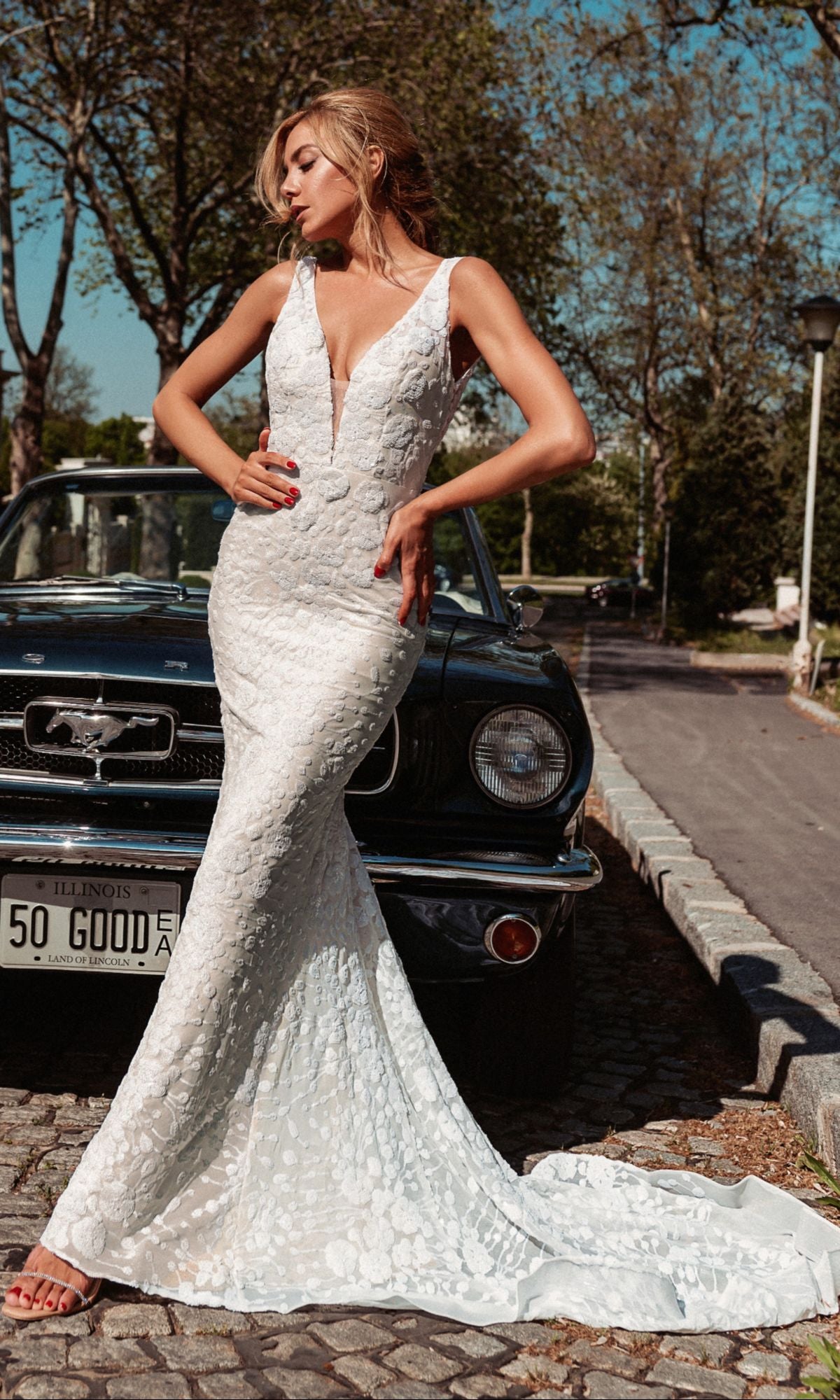 A woman wears the Long White Mermaid Bridal Gown: Tina Holly BB011 in White/Ivory, standing in front of a vintage black Ford Mustang on a cobblestone street.